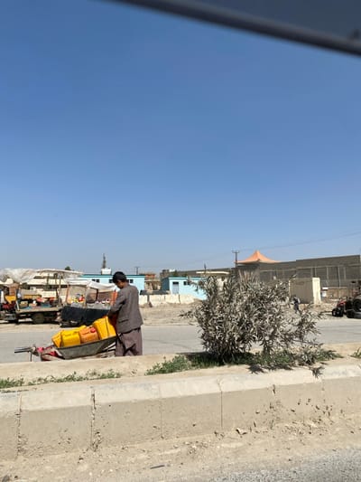 An individual, dressed in brown, appears to be attending to a cart filled with yellow containers on a roadside. The roadside scene includes a concrete curb with vegetation along the edge. A building with an orange dome roof and smaller structures are visible in the background under a clear blue sky.