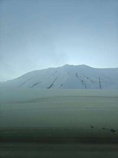 The image depicts a snow-covered landscape in what is likely the city of Ganisi, Georgia, based on the prompt. A large, snow-covered mountain dominates the background, partially obscured by haze or fog at its base. In the midground, a flat, snow-covered area is visible, punctuated by the presence of two power pylons/electrical transmission towers. The foreground features a dark, indeterminate surface, possibly a road or the interior of a vehicle, with indistinct dark shapes. The sky is overcast, contributing to the overall monochromatic and subdued tone of the scene. No people or animals are visible.
