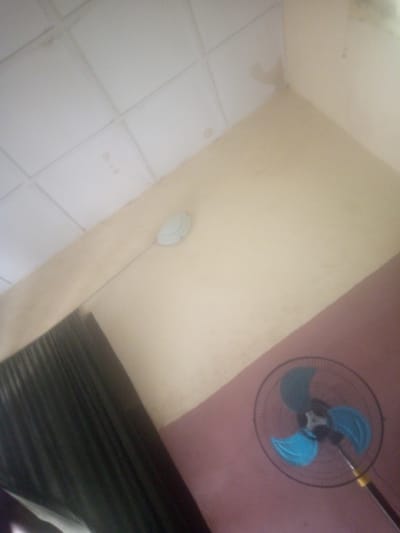 The image depicts an indoor scene with a ceiling fan and part of a standing fan visible. The ceiling appears to be made of acoustic tiles with some water stains. The walls are painted in a light yellow color, with a section transitioning to a reddish-pink hue. A dark curtain is visible on the left side of the image. The standing fan has a black grille and bright blue blades. The overall lighting suggests it is daytime, likely indoors with artificial light. The image seems to be taken from a low angle, looking upwards. There is no discernible text or specific activity taking place beyond the presence of the objects. The setting is likely a room in Jauro Bamvo, Nigeria.