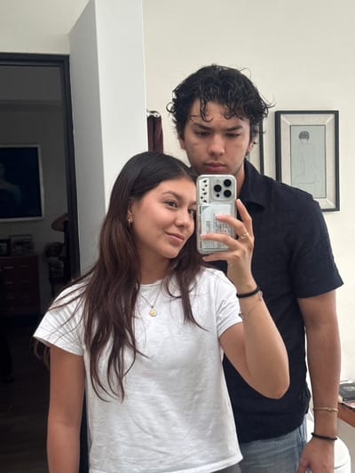 A selfie-style photograph shows two individuals, a male and a female, inside an interior space in Oaxaca City, Mexico. The female is holding a smartphone with a clear case adorned with black stars, taking the picture. She is wearing a white t-shirt and a necklace with a hexagonal pendant. Her dark brown hair falls across her shoulder. She is looking directly at the camera. The male is wearing a black shirt and is positioned behind her. He is also looking at the camera. A framed artwork hangs on the wall behind them, possibly a line drawing. The room appears to be part of a residence, with a doorway leading to another area visible in the bottom left. An additional picture hangs behind the woman and on a table. The lighting is even, with the image appearing to be taken indoors during the daytime. Both people are wearing bracelets. The man is wearing a gold one on his wrist and another is black. The woman is wearing two bracelets on her wrist as well.