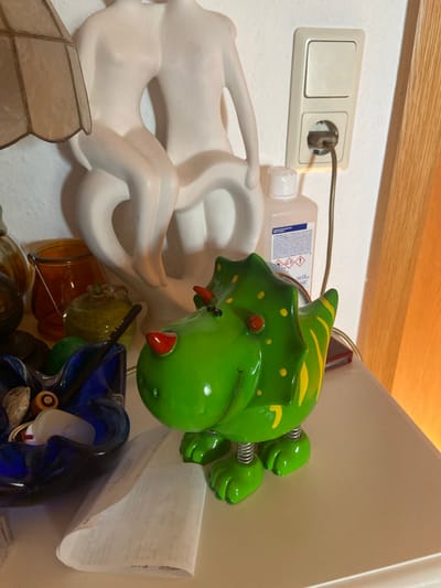 The image depicts a shelf in a room, possibly a bedroom or living area, located in Kassel, Germany. On the shelf, several items are visible: a white sculpture of two figures embracing, a lamp with a patterned shade, a bottle of hand sanitizer, a green ceramic dinosaur figurine with yellow stripes and red accents on its nose and horns, and a blue glass ashtray containing various objects, including what appear to be stones and a battery. A white electrical outlet with a plug and cord is mounted on the wall behind the sanitizer bottle. A folded piece of white paper with printed text is also on the shelf in front of the ashtray. The overall scene suggests a domestic setting with decorative objects and functional items.