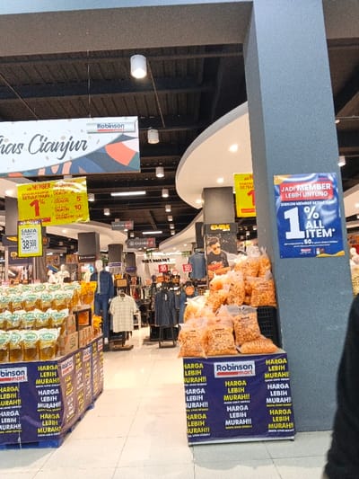 The image captures an aisle within an indoor retail store, likely a supermarket or department store, indicated by various product displays and promotional signs. The setting is brightly lit with overhead lights illuminating a black, open ceiling with exposed pipes.
On the left side of the aisle, a large display of cooking oil, specifically "SANIA" brand, is visible, priced at 38,500. Below these products are blue banners with white text repeatedly stating "HARGA LEBIH MURAH!!" (Cheaper Price!!).
To the right, a prominent grey pillar features a blue promotional sign that reads "JADI MEMBER LEBIH UNTUNG" (Become a Member, More Benefits) and "DISKON MEMBER SETIAP HARI 1% ALL ITEM" (Daily Member Discount 1% All Item). Adjacent to the pillar, a large display of packaged snacks, resembling puffed corn or cheese balls in clear plastic bags, is stacked on a stand also adorned with "HARGA LEBIH MURAH!!" banners.
In the background, clothing sections are visible with mannequins dressed in denim and casual shirts, alongside racks of apparel. A "CASHIER" sign with an arrow points towards the back, and another sign for the "EMBA" brand can be seen. Various yellow and red promotional signs are scattered throughout the store, some mentioning "DISKON MEMBER" and percentages like "1%" and "5%".
Overhead, a large white banner with colorful graphics hangs, bearing the text "Khas Cianjur" (Specialties of Cianjur) and the "Robinson mart" logo, suggesting the store's location or a focus on local products from the Cianjur region. The overall scene indicates an active retail environment with an emphasis on discounts and membership benefits.