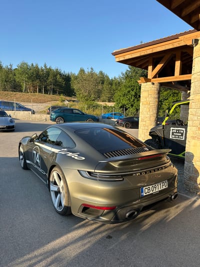 The image captures a sunny daytime scene outside a building with a wooden and stone facade, likely in Kerenite, Bulgaria. The primary focus is on a row of parked cars, with a Porsche Turbo prominently displayed in the foreground, its metallic green paint shimmering in the sunlight. The car features the word "Turbo" in white lettering along its side and a "74" graphic. A Bulgarian license plate, "CB 0911 PO," is clearly visible.

Behind the Porsche, another car, a dark grey sedan, is partially visible, and further back, a dark green SUV is parked. In the background, there's a black ATV with a yellow frame, displaying Cyrillic text that reads "АТВ И УТВ РАЗХОДКИ" and a phone number, suggesting it might be available for rental or tours. The scene is set against a backdrop of lush green trees and a clear blue sky, indicating fair weather. The shadows cast by the building and trees suggest it's likely late afternoon. The overall impression is one of a leisure or travel destination, possibly a resort or a scenic spot.