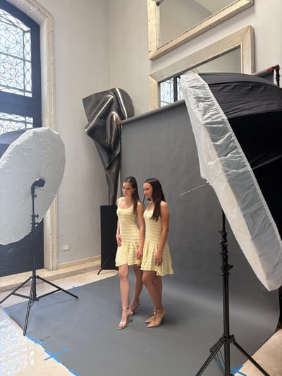 The specific location within Monterrey, Mexico, is not visually discernible from the image. The scene depicts an indoor photography session set up in an ornate room. Two young females stand on a seamless grey backdrop that extends from the wall to the patterned light-colored tile floor, secured with blue tape. Both females wear identical or very similar pale yellow, sleeveless, knee-length dresses with a horizontally ribbed texture and ruffled hems. The female on the left wears silver strappy high heels and looks slightly to her left. The female on the right wears studded nude-toned high heels and looks towards the female on the left with a subtle smile.
Professional photography equipment frames the subjects: a large round softbox on a stand is positioned to the left, and a large rectangular strip softbox on a stand is positioned to the right. In the background, behind the grey backdrop and to the left, a large, dark, abstract metallic sculpture is displayed on a pedestal. Above the backdrop, on a light-colored wall, two framed mirrors with distressed gold-toned frames are mounted. To the far left, a tall, dark-framed arched window with intricate white grilles is visible, suggesting an exterior view.