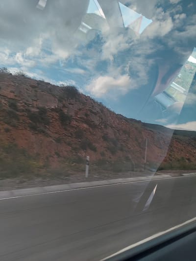 The image, taken from inside a vehicle and rotated counter-clockwise, presents a layered view of an outdoor scene and interior reflections under bright daylight.

In the background, a rugged, reddish-brown mountain or cliff face, textured with rocks and sparse green vegetation, dominates the left side. To its right, a gray paved road with white markings (both solid and dashed) indicates a highway. A small white marker or post is faintly visible near the mountain edge.

Superimposed are reflections from the vehicle's interior: a vast expanse of blue sky with white, fluffy clouds, particularly prominent in the upper and central parts of the image. Other reflections include a reddish-brown shape, possibly part of a car seat or dashboard, in the upper left, and a triangular reflection with a red border in the mid-left. While its specific symbol is not clear, it appears to be a warning sign. Faint outlines that could be a person's head or arm are also reflected.

The scene captures a moment of travel through a mountainous landscape, consistent with the provided context of Tigranashen, Armenia. The weather appears clear and bright. No discernible text is visible on any signs or objects, apart from the inferred type of the triangular warning sign.