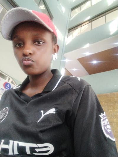 A medium close-up portrait captures a young woman, likely in her late teens or early twenties, posing indoors in what appears to be a modern building in Sheywe ward, Kenya. She has dark skin and short dark hair, partially covered by a red and grey baseball cap worn backward. Her lips are slightly pursed in a common selfie pose, and she looks directly at the camera with a neutral expression.
She is dressed in a black collared t-shirt with white trim on the collar. The shirt features a white Puma logo (a leaping cougar) on the chest, and below it, partially visible white text that reads "HITE". On her right sleeve, a distinct purple and white Premier League logo is visible, indicating an affiliation or interest in the English football league. A light-colored, possibly white, abstract earring adorns her right ear.
The background suggests an indoor setting with contemporary architecture, featuring light blue-green walls and multiple large windows or glass panels that reflect bright natural light. These reflections indicate a well-lit space, likely during the daytime. A blurry blue and white sign can be faintly seen in the distance behind her left shoulder.