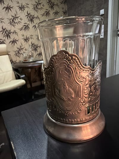 A close-up shot shows a glass in an ornate metal holder, possibly a Soviet-era tea glass holder, resting on a dark, textured surface. The glass has vertical ribbed detailing and is transparent, revealing some contents within, though not clearly discernible. The metal holder is detailed with embossed patterns, including what appears to be a building and foliage, and has a slightly aged, coppery patina. The background features a wall with a wallpaper pattern of dark leaves against a lighter background. To the left, a portion of a light-colored, upholstered chair is visible, and further back, a dark wooden table with a circular top. A bright light source is reflecting off the metal holder and the glass. The scene is indoors, likely in a residential or formal setting. The image does not provide information about the city of Bissendorf, Germany.