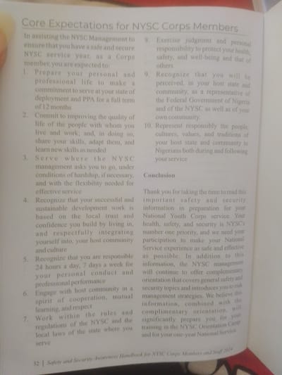The image is a close-up, slightly angled photograph of an open book. The text on the page is printed in a light gray font against a white background. The book appears to be a handbook, as indicated by the title "Core Expectations for NYSC Corps Members" at the top of the page and a footer that reads "Safety and Security Awareness Handbook for NYSC Corps Members and Staff 2024". The content is a list of numbered expectations and a section titled "Conclusion". There are no people, objects, or environmental features visible beyond the book itself. The specific location in Magajin Gari, Nigeria, cannot be determined from the provided image.