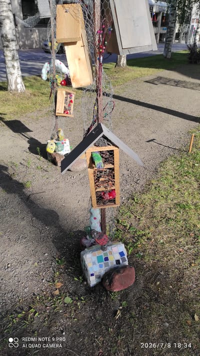 This is a photograph taken outdoors on a sunny day in Iisalmi, Finland. The subject is a homemade insect hotel constructed from wood and wire mesh, attached to a metal pole. The insect hotel has several compartments filled with natural materials like twigs and leaves, and some compartments contain colorful stuffed objects. At the base of the insect hotel, there is a mosaic-covered block and a large, reddish-brown rock. The background shows a paved pathway, grass, trees, and a building. The photograph has a timestamp in the bottom right corner indicating "2026/7/8 18:34" and a watermark in the bottom left corner reading "REDMI NOTE 9 PRO AI QUAD CAMERA".