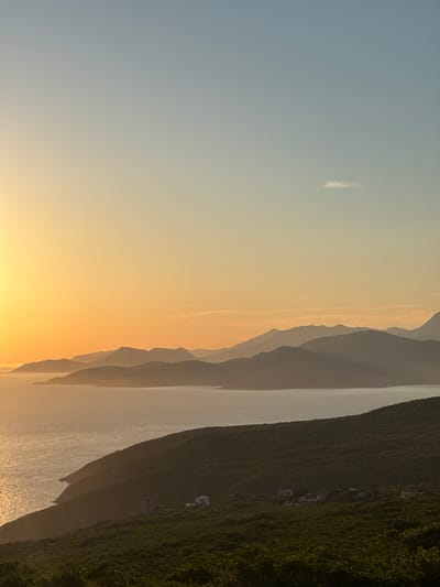 An aerial vertical shot captures a serene sunset over Kovačko Polje, Montenegro. The sun, a bright golden orb, sets ablaze the sky with hues of yellow and orange. A gentle sea laps at the shore, its surface shimmering with the sun's golden light. In the foreground, a lush green hill is dotted with a few sparsely placed houses, their architecture simple and functional. The background is dominated by a series of undulating mountains, their peaks fading into the hazy horizon, creating a sense of depth and grandeur. The sky transitions from a warm yellow at the horizon to a soft blue at the zenith, with a single wisp of cloud drifting lazily across the expanse. The scene evokes a sense of peace and tranquility, a perfect encapsulation of a Mediterranean evening.