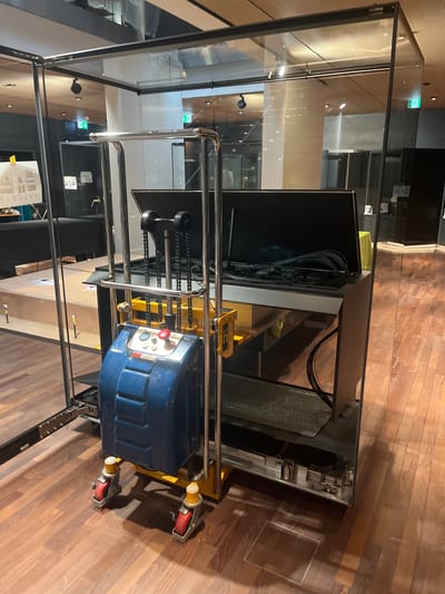 The image shows a piece of scientific equipment enclosed in a glass casing. The equipment is a metal frame with a blue and yellow component at the bottom, featuring a red button and some dials. There are chains hanging from the top of the frame, connected to the blue component. The casing is a large, rectangular structure with clear glass panels supported by a metal frame. The floor is made of polished wooden planks. In the background, there is a large pillar, ceiling lights, and what appears to be an exit sign with a green running figure. The scene is indoors, possibly in a museum or exhibition space. The location is the CERN facility in Cologny, Switzerland.