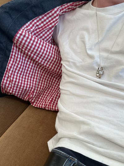 The scene depicts a close-up, top-down view of an individual's upper torso and lap, situated in Clichy, France. The subject wears a white short-sleeved t-shirt, blue denim jeans, and a black belt. A silver-colored chain necklace with a two-part pendant, each section appearing encrusted with clear stones, hangs over the t-shirt. An outer garment, possibly a jacket, is worn open on the left, revealing a distinctive red and white gingham pattern lining. The exterior of this garment is a dark blue, likely denim. The person is seated or reclining on a brown, upholstered surface, possibly a sofa or armchair, which occupies the lower left and bottom portions of the frame. No specific actions or interactions are discernible beyond the person's static posture.