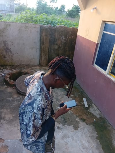 A medium shot captures a young man with dreadlocks, wearing a camouflage jacket, standing outdoors and looking down at his smartphone. He is positioned on a rough, wet concrete surface, with a concrete wall to his left and a painted building to his right. The wall is textured and shows signs of age, with visible cracks and stains. Behind the wall, lush green vegetation and trees are visible, suggesting a natural or semi-urban environment. To the man's left, a circular well with a stone cover is partially visible, indicating the presence of a water source or a drain system. The building on the right has a two-toned paint scheme of beige and dark pink, with a blue-framed window. An outdoor light fixture is mounted on the wall of the building, above the window. The lighting suggests it is either early morning or late afternoon, with soft natural light illuminating the scene. The overall environment appears to be a residential area or a less developed part of a town in Ukukwa, Nigeria, given the context. The man is holding a black smartphone in his right hand, with his left hand partially visible. White earbuds with a wire are connected to the phone. The ground is damp and reflective, with some debris scattered around.