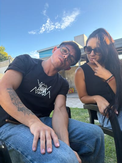 Two individuals, a male and a female, are seated outdoors in Quilmes Oeste, Argentina, under a clear blue sky with scattered clouds. The scene is illuminated by strong daylight, indicating late afternoon.
The male, positioned on the left, is a young adult with short dark hair, wearing black-framed eyeglasses, a black short-sleeved t-shirt with a white "DMIA" graphic, and light blue jeans. His head is tilted to his left, and he gazes directly at the camera with a slight pout. A floral tattoo is visible on his left forearm, and a thin silver chain is around his neck. His left hand rests on his thigh; a slender, dark cylindrical object is partially visible beneath it.
The female, positioned on the right, is a young adult with long dark hair cascading over her right shoulder. She wears dark sunglasses with light brown frames, a black sleeveless button-front top, and light blue jeans. Her right index finger is pressed against her lower lip as she looks slightly to her left. Multiple thin bracelets adorn her right wrist. Her left hand rests on the armrest of her chair, grasping a section of her hair.
Both individuals are seated in dark-colored outdoor chairs, only partially visible. The immediate foreground features green grass, transitioning to a paved or tiled area in the mid-ground. The background includes light-colored concrete walls or fences, a modern building with a prominent horizontal light blue section, and distant, indistinct reddish-yellow objects.