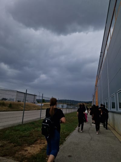 A group of individuals, most wearing dark clothing, are walking along a paved path beside a large, modern, grey building with orange trim and a row of windows. The path is bordered by a grassy area and a chain-link fence. In the distance, industrial buildings are visible behind the fence. The sky is heavily overcast with dark, dramatic clouds, suggesting an impending storm. The scene appears to be an industrial or commercial area, possibly on the outskirts of the city of Bahovitsa, Bulgaria. One individual in the foreground, facing away from the camera, wears a black t-shirt, blue jeans, and a black backpack. The other individuals are further down the path, walking in the same direction. The overall atmosphere is subdued and somewhat ominous due to the weather conditions.