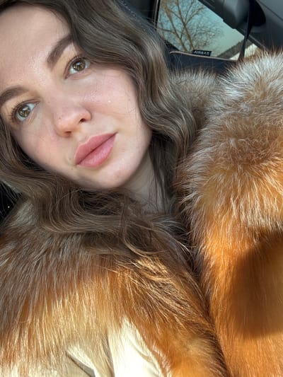 An adult female subject is depicted in a close-up selfie taken from within a vehicle in Nazhitsy, Russia. Her upper body is visible, including her face, wavy light brown hair, and shoulders. She has fair skin, greenish-brown eyes directed slightly upwards and to her left, and wears light pink lipstick. A small, white blemish or mark is present on her right cheek. She is dressed in a prominent reddish-brown fur collar, likely from a coat or jacket, over a lighter-colored garment. The vehicle's interior features dark trim, and a side window in the upper right background reveals bare tree branches against a bright sky. An "AIRBAG" label is visible on the vehicle's interior panel near the window. Strong natural light illuminates the subject, particularly highlighting the fur.