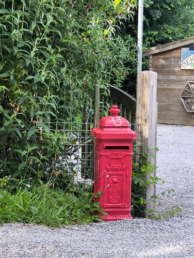 A vibrant red, ornate post box, featuring traditional decorative elements, stands on the left side of a gravel pathway. The number "13" is painted in white on its domed top, and a mail slot is visible on its front face. Lush green foliage, appearing as dense bushes or a hedge, forms a natural backdrop behind and to the left of the post box, partially obscuring a metal wire fence or trellis.
To the right of the post box, a sturdy light-colored wooden post is positioned, with the gravel path extending into the background on the right. Behind this post, a rustic wooden shed with horizontal planks is visible. Attached to the shed's side is a colorful mosaic artwork that partially spells "HEK" and features a circular pattern. Below the mosaic, a hexagonal insect hotel, constructed with various natural materials, is also mounted on the shed.
The ground in the foreground and extending to the right consists of a gravel surface, with patches of green grass and weeds growing along its edges. The scene is illuminated by natural daylight, indicating daytime. The setting suggests an outdoor, possibly garden or semi-rural, environment, in Alkmaar, Netherlands. No specific activity or individuals are present.