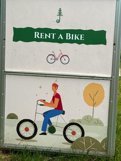 The image shows a two-panel sign, likely for a bike rental service, placed outdoors. The left panel features a stylized green tree silhouette and the text "RENT A BIKE" in large white letters against a white background, bordered by a green, irregularly shaped block. To the right of the text, there is a simple illustration of a red bicycle.

The right panel depicts a cartoonish person in a red shirt and blue pants riding a bicycle. The rider is leaning forward, appearing to pedal. The background of this panel includes a yellow sun-like circle and stylized green trees with leaves, suggesting an outdoor, possibly park-like setting. There are also some small starburst shapes on the right panel.

The sign is framed by a metal border, and the overall image suggests it is situated outdoors, as evidenced by the grass visible around the edges of the sign and the natural lighting. The weather appears to be clear, and the time of day is likely daytime, given the bright illumination. The cheerful style of the illustrations suggests a lighthearted and welcoming atmosphere. No specific emotions are overtly displayed by the figures, but the activity of cycling in a natural setting evokes a sense of recreation and enjoyment. There are no explicit location cues beyond the provided context of Dilijan, Armenia.