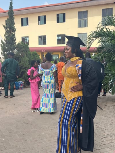 The image depicts a graduation scene in Madina, Ghana. The primary subject is a woman positioned slightly right of center, wearing a black graduation cap with a tassel, a black gown, and a colorful striped stole. Beneath the gown, she wears a yellow, beaded, sweetheart-neckline top and blue, white, and yellow vertically striped wide-legged trousers. Her left hand rests on her hip as she smiles and gazes towards the left.
To her immediate left, a woman is seen from the rear, wearing a long, horizontally patterned dress composed of light blue, yellow, pink, and white rectangular blocks, and carrying a small black handbag. Next to this woman, a young girl in a bright pink peplum top and matching trousers, wearing white low-heeled slip-on sandals, faces left. Further back on the left, a man in a dark green traditional long-sleeved tunic and trousers stands. Additional blurred figures are visible in the background, including one in an orange outfit.
The background features a two-story, light yellow building with a reddish-brown sloped roof and a dark brown base trim. It has multiple rectangular windows with dark frames. Various green trees and shrubs are present, including a tall, slender conifer on the far left and a broad-leafed plant on the right, partially obscuring a silver photographic umbrella/reflector. The ground is paved with light-colored interlocking concrete blocks. A stack of bottled water is visible on the far left.