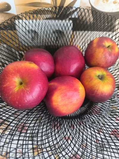 The image shows a black wire fruit basket filled with six apples. The apples are red and yellow, with some showing a gradient of color. The basket is sitting on a surface with a floral pattern. The background is blurred, but appears to be a wall with a textured pattern. The lighting suggests it is daytime, possibly indoors. No people, text, or specific activities are visible. The location context is Bender, Moldova.