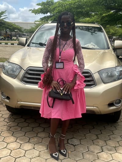 A young Black woman stands in front of a gold SUV. She is wearing a pink dress with lace sleeves and a ruffled hem, black high heels with heart-shaped embellishments, and a black handbag. She has long braided hair and is wearing sunglasses. The vehicle is parked on a paved area in what appears to be a public space, possibly an event venue or parking lot in Itoga, Nigeria. Trees and other vehicles are visible in the background, suggesting an outdoor setting within the city. The overall impression is of a formal occasion or outing.