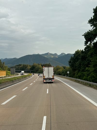 The image depicts a highway scene with mountainous terrain and trees in the background. A white truck with a trailer is the prominent object in the foreground, traveling on the road. Several other vehicles, including cars and smaller trucks, are also visible on the highway. The road is paved and has lane markings, suggesting it's a multi-lane highway. A guardrail separates the road from the embankment on the left, which is covered in grass and trees.
The scene appears to be set during the daytime, with an overcast sky. The weather seems to be cloudy, as indicated by the diffused light and the absence of strong shadows. The mountains in the background are covered with trees and have a rugged, natural appearance. The presence of a highway winding through this natural landscape suggests a scenic route, possibly in a mountainous or hilly region. The crops indicate the location is Raubling, Germany.
Visible text on the back of the white truck's trailer includes "SCHMITZ" and possibly other smaller markings or logos. There are also white dashed lines on the road, indicating lane separation. A circular sign is visible on a pole near the guardrail, but its text is not legible. The overall impression is that of a journey on a highway, likely during a cloudy day, passing through a picturesque natural environment in Germany.