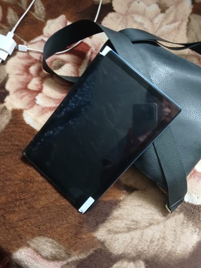 This is a close-up, high-angle shot of a black tablet placed on a brown and beige patterned blanket. A black strap from a bag is visible draped over the tablet and extends to the right. To the upper left of the tablet, a white USB power adapter is connected to a white cable. The tablet's screen is dark and shows some smudges. The scene is indoors, and the objects are arranged haphazardly on the blanket, suggesting a casual or temporary setting. There is no indication of the specific location within Hlushky, Ukraine.