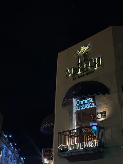 At night, a sign for the "MAKECH BY CASA MAYA" restaurant stands out. The sign is in yellow lighting. Adjacent to it, a blue neon sign reads "Comida Yucateca". Above, the restaurant has a small balcony with stairs leading up to it, marked by a neon sign that reads "ENTR DALE MAKECH" with an arrow pointing to the entrance. In the distance, illuminated architectural details are visible. This photograph was taken in Mérida, Mexico, capturing the vibrancy of the restaurant's signage in the darkness.