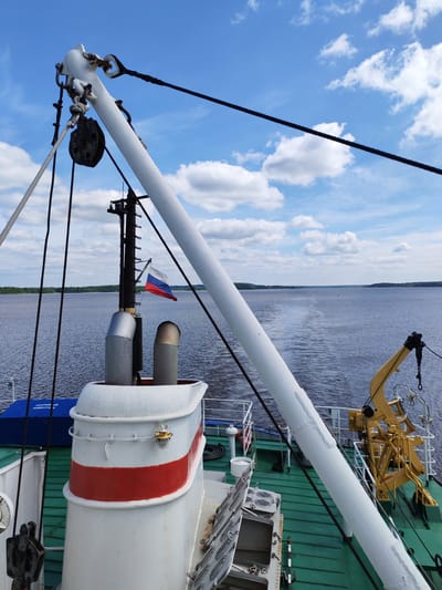 A partial view from the deck of a boat shows the top of a smokestack with a horizontal red stripe, two metal exhaust pipes, and a black mast flying the Russian flag. A white boom stretches from the top left toward the lower right of the frame, supported by black and white cables and a pulley system. In the distance, a wide body of water reflects the blue sky dotted with white clouds, and a faint wake suggests the boat is moving. The deck of the boat is green. This scene is likely somewhere in Russia. It is unknown if Pruzhinino is the current location.