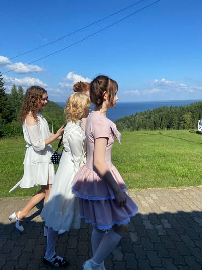 In Galevo, Russia, a group of four young women are walking along a paved pathway with a scenic backdrop. The woman on the left wears a white off-the-shoulder dress and white heels, carrying a black and white polka-dot purse. Next to her, a woman in a white ruffled dress is visible. The third woman from the left is wearing a pink dress with white lace trim and white thigh-high socks. The fourth woman, partially visible on the right, also appears to be wearing a pink outfit. They are walking towards the right side of the frame. The environment features a lush green grassy hill, a dense forest, and a body of water in the distance under a clear blue sky with scattered clouds. A white structure is visible on the right side of the image, situated on the edge of the hill.
