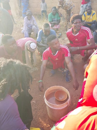 The image captures an outdoor gathering of approximately 12-15 individuals, predominantly male, in Vodni, Nigeria, on reddish-brown earth. Most people are squatting or crouching in a loose semi-circle. In the immediate foreground, a white plastic bucket containing a reddish-brown liquid and a brown, bowl-shaped gourd rests on the ground. A man in a red Adidas t-shirt and shorts, wearing a green wristband, squats directly behind the bucket, looking towards the right. To his right, another man in a similar red Adidas outfit with a brown knitted cap also squats. To the left of the central figure, a man in a maroon shirt, wearing a white and brown patterned cap with a light cloth draped over his head, leans forward.

Various other individuals populate the midground and background. These include an older man in light-colored traditional attire, squatting and smiling; a woman in a green t-shirt and patterned skirt, squatting; and a man in a yellow t-shirt, also squatting. A young boy in a light hoodie stands in the distant background, near a structure with mud or unpainted brick walls. An old tire is visible leaning against a wall on the far left. The group appears to be engaged in a communal activity, potentially involving the shared liquid in the bucket.