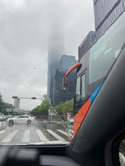 The image depicts a view from inside a vehicle, looking out onto a wet city street in Futian District, China. Raindrops are visible on the windshield, distorting the view of the scene outside. Tall, modern skyscrapers dominate the background, their upper sections obscured by heavy fog or clouds. Traffic lights are red, indicating a stop. A white sedan is positioned on the wet road, in front of a pedestrian crossing. To the right, a portion of a large bus is visible, with an orange side mirror. The sky is overcast and grey. The overall environment suggests a rainy day in a dense urban setting.