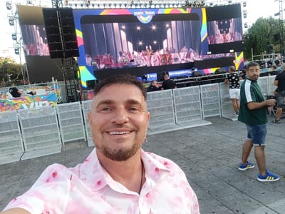 A man in a pink floral shirt is taking a selfie in front of a large outdoor screen showing a performance. The screen displays "Eurovision Palomas Festival." Several people are standing behind a metal fence, watching the performance. One man, in a green shirt and jean shorts, is visible on the right side of the frame. The ground is paved with grey stone tiles. The setting is likely a public square or event space in San Bartolomé de Tirajana, Spain, given the festival mentioned.