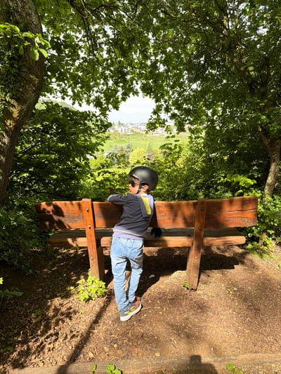 A young person wearing a black helmet, sunglasses, a long-sleeved navy blue shirt, light blue jeans, and multicolored sneakers is lying prone on a weathered wooden bench. The bench is located in a wooded area in Pétange, Luxembourg. A view of the town is visible in the distance, framed by trees and foliage. The ground surrounding the bench is covered in mulch, and dappled sunlight shines through the trees.