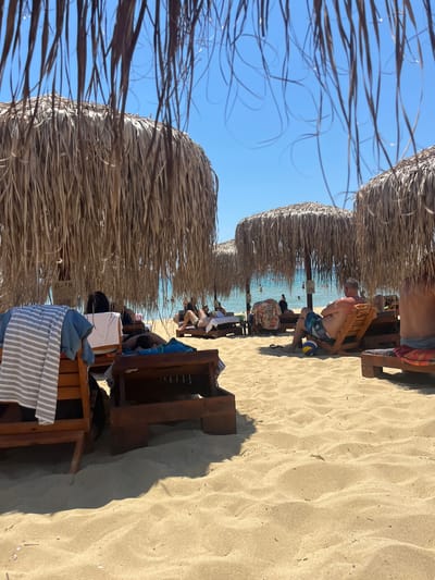 This media file captures a sun-drenched beach scene in Nea Peramos, Greece. The image is oriented vertically. In the foreground, the dried fronds of thatched umbrellas stretch across the frame, creating a natural canopy. Beneath these umbrellas, rows of wooden lounge chairs are set up on the golden sand.
Several people are visible relaxing on the chairs. One individual, a man with a patterned swim trunk, is seated, with a volleyball at his feet. Further back, other beachgoers are seated or walking, with the turquoise water of the sea visible in the distance, reflecting the clear blue sky. The lighting suggests it is daytime, likely late morning or early afternoon, given the strong sunlight and pronounced shadows cast by the umbrellas and chairs. The weather appears to be clear and warm, typical for a beach day.
In the mid-ground, a wooden structure appears to be a beach bar or service station, with some signage. One sign, partially visible, displays "B-8". Towels, one striped and one blue, are draped over a lounge chair in the lower right portion of the frame. The overall mood is one of leisure and relaxation, with people enjoying a day at the beach.