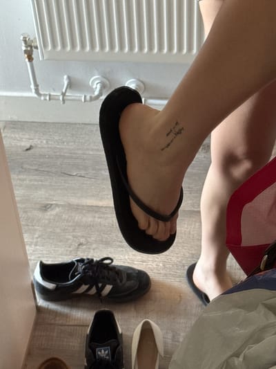 The image displays a close-up, low-angle view of a person's legs and feet in an indoor setting in Hull, United Kingdom. One foot, adorned with a black flip-flop, is prominent in the foreground and slightly elevated. A small, calligraphic script tattoo is visible on the top of this foot and ankle, which reads "living my life in peace". The person's other leg and foot, also in a black flip-flop, are partially visible in the background, along with a glimpse of a red and white striped garment worn by the individual.

The floor is made of light-colored wooden planks, featuring a visible grain. To the left, a white wall-mounted radiator with its associated piping is clearly visible. Scattered on the wooden floor are several other pairs of shoes: a pair of black Adidas Samba sneakers with white stripes and laces, light-colored flat shoes or slippers, and a partially visible brown shoe or sandal on the far right. A light-colored, possibly fabric bag or object, is also seen near the person's other leg. The scene is well-lit, suggesting either natural daylight or bright indoor illumination. The image focuses on personal details like the tattoo and footwear within a casual home environment.