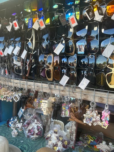 A close-up, eye-level shot captures a stall displaying a variety of sunglasses and keychains. The sunglasses, predominantly black and featuring tinted lenses, are arranged in rows on a metal rack. Some are adorned with price tags and brand labels such as "OAKLEY," "RB Sport," and "RODEO." Below the sunglasses, numerous keychains hang from hooks. These keychains are diverse, featuring shell decorations, wooden carvings, miniature figures, and colorful plastic charms. The stall is set against a backdrop of what appears to be a market or outdoor vendor setting. In the foreground, the table is covered with a teal-patterned fabric, upon which several packaged items and decorative shell arrangements are displayed. The overall scene suggests a commercial display of accessories.