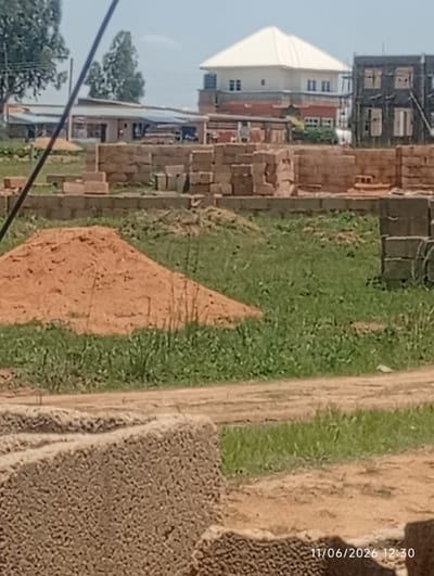 This image depicts a construction site in the city of DU, Nigeria, on June 11, 2026, at 12:30 PM. The foreground shows a pile of bricks and a mound of sand, suggesting ongoing construction work. In the middle ground, a partially constructed building with brick walls is visible. The background features a larger building with a white roof, possibly a residential house, and another building under construction with exposed windows and scaffolding. Trees and vegetation are present, indicating an outdoor setting. The sky is clear, suggesting fair weather. No people are visible in the image.