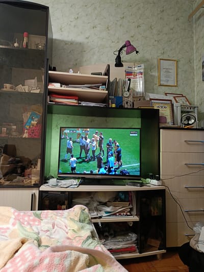 This is a full shot image of a living room with a television in the center displaying a soccer match. The room has a cluttered but lived-in feel. To the left, a tall, dark-colored cabinet with glass doors contains various items on its shelves, including what appear to be bottles, books, and decorative objects. To the right of the television, a dresser with light-colored drawers is visible. The walls are covered in a light-colored wallpaper with a subtle marbled pattern. Above the television, on a shelf, are several framed documents and awards, including a gold-framed certificate. A small pink desk lamp is also attached to this shelf. The television rests on a dark media console, which is also filled with papers and other miscellaneous items. In the foreground, a soft, patterned blanket is draped over what appears to be a sofa or chair. The lighting suggests it is either daytime or evening with indoor lights on. The presence of specific Russian writing on some of the documents and awards suggests a Russian context, potentially Arkhangelsk, Russia, based on the prompt.
