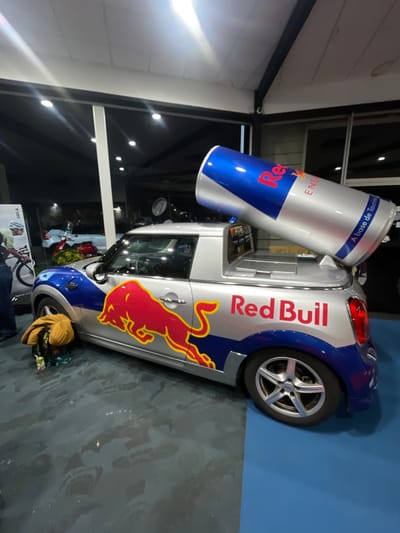 A silver car, heavily branded with "Red Bull" logos, stands on a gray floor against a blue wall. The car's design includes a large Red Bull can mounted on the back, mimicking a truck bed. A red bull graphic adorns the side of the car. In the background, a bright, light-filled room is visible through the glass, with hints of other vehicles and displays. A person's leg is visible in the foreground.