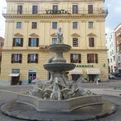 The image depicts the Fontana del Calamo (also known as delle Tredici Cannelle) in Ancona, Italy. In the foreground, a multi-tiered stone fountain features a cherubic figure at its apex, from which water cascades into a large central basin. Below this, seven sculpted winged horses (pegasi) are positioned in the lowest basin, spouting water from their mouths. The fountain is set on a light-colored paved square.

Behind the fountain stands a prominent, four-story, pale yellow building with ornate architectural details. Its facade features numerous rectangular windows, mostly fitted with dark brown wooden shutters, some open, some closed. The uppermost stories have iron-railed balconies, one with visible greenery. The ground floor of this building houses several businesses: "GAVETTI" and "M.E.D.I.A." are visible on the left, while "FARMACIA" (pharmacy) occupies a larger section on the right, distinguished by green lettering and a green cross symbol. The pharmacy has two white awnings labeled "FARMACIA DEI RAGAZZI".

Two individuals are visible near the pharmacy entrance, one wearing a light-colored top and dark pants, the other a light blue top and white pants. To the right of the main building, a street slopes uphill, with several parked cars lining its side. Other residential buildings, including one with light green shutters and another with brick-colored facades, are visible further along this street. The sky above is partially cloudy.