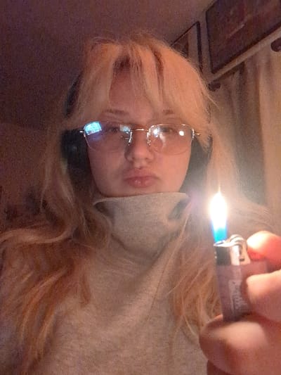 The image captures a young individual, likely female, with blonde hair and bangs, wearing round-frame glasses and black over-ear headphones. She is dressed in a light-colored turtleneck sweater. Her lips are pursed, and she gazes directly at the camera. In the foreground, her right hand holds a lit disposable lighter, emitting a bright blue flame. Reflections of a digital screen are visible on the lenses of her glasses. The background reveals an indoor setting with low light, featuring a light-colored curtain and a framed object on a wall. The scene is located in Area D (Elaho/Garibaldi), Canada.