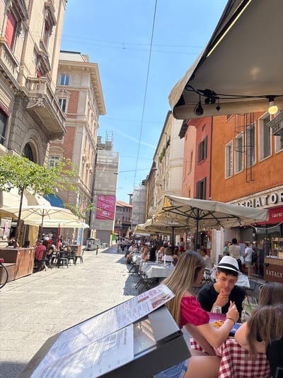 The image captures a lively street scene in Bologna, Italy, with people dining outdoors at numerous tables lining the pedestrian street. The architecture of the buildings is characterized by a mix of orange, red, and beige facades with varied window designs and detailed molding. A prominent sign for "EATALY" is visible on one of the buildings.
People are seated at the tables, engaged in conversations and eating. A young man wearing a hat and a person in a red and white checkered shirt are noticeable at one of the tables. A restaurant menu is prominently displayed in the foreground.
White parasols provide shade for the diners, and a few small trees are interspersed along the street. Overhead wires are visible against the clear blue sky. The ground is paved with light-colored stones. The overall impression is of a bustling and sunny day in a historic Italian city.