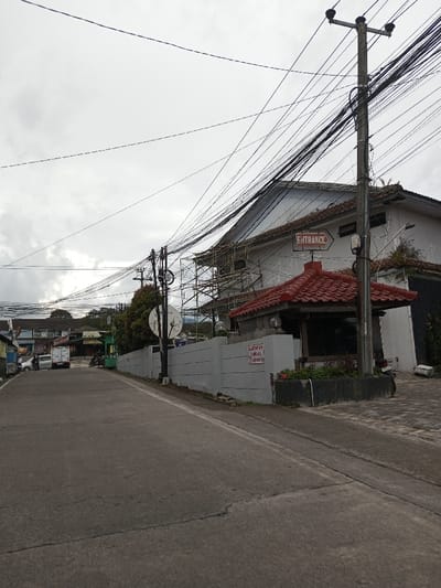 This image depicts a street view in Melakudikadu, India, on an overcast day. A paved road, exhibiting minor cracking, occupies the lower and central portions of the frame, ascending slightly into the background.
On the right side of the road, a low, light grey concrete wall runs parallel to the thoroughfare. Behind this wall, a structure with a distinct red, tiered tiled roof is visible, appearing to serve as an entrance or small kiosk. Above this, a two-story white building, displaying signs of wear, features brown roof tiles and is partially obscured by metal scaffolding erected against its facade, suggesting ongoing construction or renovation. A red sign with white lettering, reading "ENTRANCE" and featuring a rightward-pointing arrow, is affixed to this building. A prominent dark grey utility pole, laden with numerous black electrical and communication wires, stands on the right side, extending upwards and across the frame. Another smaller utility pole is visible further down the street to the left. A white satellite dish is mounted on a pole behind the grey wall. A white sign with red text (in a local language) is attached to the grey wall.
On the left side of the road, various single and two-story buildings, appearing to be commercial establishments or residences, line the street. A white light-duty truck is parked on the left side of the road, further down. A motorcycle is also visible parked near a green-fronted establishment. Distant vegetation and additional structures are visible further down the street on both sides. The sky is uniformly grey and overcast, indicating a cloudy weather condition. No specific individuals are clearly identifiable or engaged in distinct actions within the visible foreground.