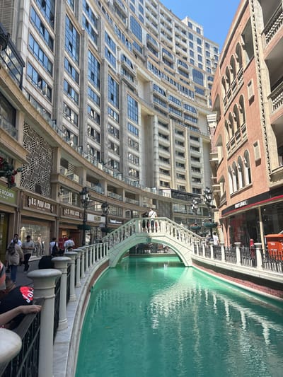 This image depicts a modern urban scene in Esenler, Turkey, characterized by a turquoise-colored canal flowing through a pedestrianized shopping and leisure complex.

On the left side of the canal, a multi-story building, light beige in color, rises prominently. It features numerous rectangular, dark-paned windows, some of which are partially open, reflecting the blue sky. Vertical decorative elements with a ribbed texture flank sections of the windows. The lower levels exhibit a patterned, geometric lattice design above glass shopfronts. Visible businesses include "Yves Rocher" and multiple "U.S. POLO ASSN." stores. A curved architectural section is apparent on the upper floors.

Opposite, on the right side of the canal, stands a multi-story building with a reddish-brown, textured facade. This building features rows of arched windows with balustrades on its upper levels, suggesting a more classical or Mediterranean architectural style. The ground floor houses retail spaces, with a red utility box or fire hydrant positioned near the far right edge of the frame.

A white, arched bridge with ornate balustrades, consisting of white pillars and black railings, spans the canal in the mid-ground, connecting the two sides. The canal water is a striking turquoise or teal color, showing ripples and reflections of the surrounding structures. The canal edges are lined with stone, with a horizontal band of reddish brick-like detailing just above the water level on the right bank.

Numerous pedestrians, casually dressed, are observed walking along the promenades flanking the canal and on the bridge. Individuals are seen in various postures, walking, standing, and observing the surroundings. Ornate black street lamps with multiple luminaires and hanging planters are spaced along the canal banks. The scene is illuminated by bright daylight under a clear blue sky, indicating fair weather conditions.