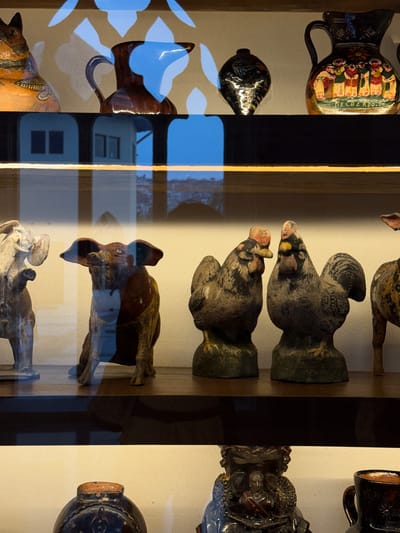 The image shows a display case, likely in a museum or gallery, possibly in San Pedro Garza García, Mexico. The case contains a variety of ceramic and sculpted objects.
On the left, there are at least two jugs or pitchers with handles. One is decorated with colorful patterns and possibly some writing, while the other is dark, possibly black. There is also a dark, teardrop-shaped object.
In the center, there are several sculptures of what appear to be birds, perhaps chickens or roosters. They are made of a dark, weathered material. There is also a sculpture of a quadrupedal animal, possibly a bull. At the bottom, there is a white sculpture of a human figure.
On the right, there is another jug or jar with a dark, patterned surface and what seems to be a sculpted figure. The background of the display case is a light tan or cream color. A window reflection is visible.