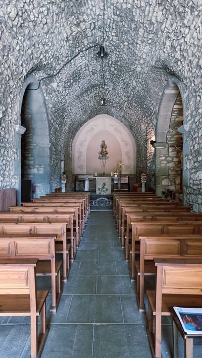 This is a full-shot, eye-level, interior, vertical photograph of the church of Santa Maria de Susqueda, in Susqueda, Spain. The church has a vaulted ceiling made of rough-hewn stones and its walls are also constructed from stone. Rows of simple wooden pews fill the nave, facing an altar at the far end. The altar area features a central focal point with a statue of the Virgin Mary, adorned with a rosary, and flanked by smaller statues and decorative elements. The floor is tiled with large, dark gray tiles. Two pendant lights hang from the ceiling. On the right side, near the front, a small table holds a magazine with the title "Full Diocesana." The overall impression is one of austerity and simplicity.
