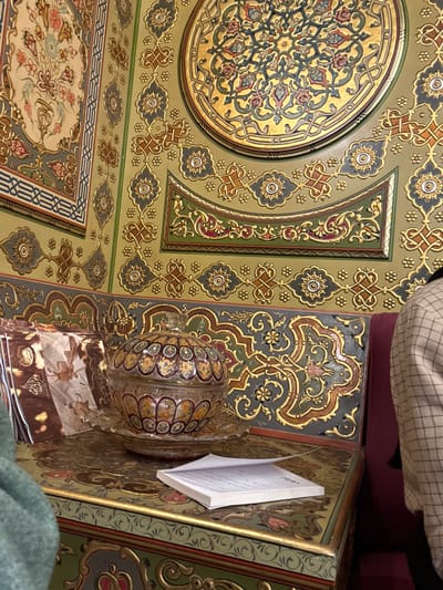 An ornately decorated ceiling and table display intricate patterns of gold, green, and gray. A circular medallion is central, surrounded by floral motifs and geometric designs. A glass bowl sits on a stack of patterned napkins. A notebook with open pages rests on the table's edge. Parts of people and clothing are visible in the background and foreground, suggesting a personal setting.