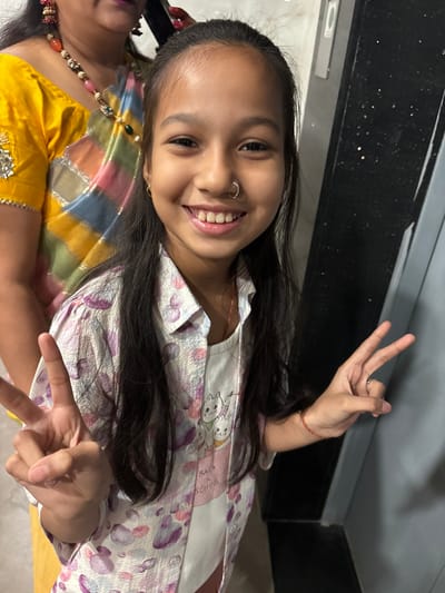 A young girl with dark hair and a nose ring smiles, making a peace sign with both hands. She wears a white patterned pajama top. A woman partially visible in the bottom left corner wears a yellow and multicolored sari with ornate jewelry. A dark, glossy countertop is in the background. The image is rotated counter-clockwise.