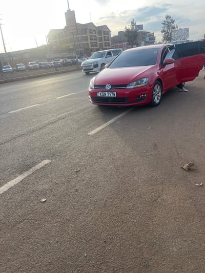 This image depicts an outdoor street scene, likely in Gitothua ward, Kenya, with a strong focus on a red Volkswagen Golf car. The car is prominently parked on an asphalt surface, angled towards the right of the frame. Its front license plate, "KDN 797W," is clearly visible, along with the Volkswagen emblem. One of the car's doors is open, and a person's legs, clad in light trousers and dark shoes, are partially visible near the opening, suggesting someone is either entering or exiting the vehicle.
In the background and extending along the left side of the frame, several other cars and a white van are parked in a line. Beyond these vehicles, multi-story buildings with varied facades line what appears to be a street, contributing to an urban or semi-urban setting. The sky is bright on the left, indicating either early morning or late afternoon, with the sun creating a strong glare and suggesting clear weather. The ground is paved with asphalt, featuring white painted lines that denote parking spots or road boundaries. Scattered small pieces of debris, including a dried leaf, are visible on the asphalt. The photograph is taken with a tilted horizon.
